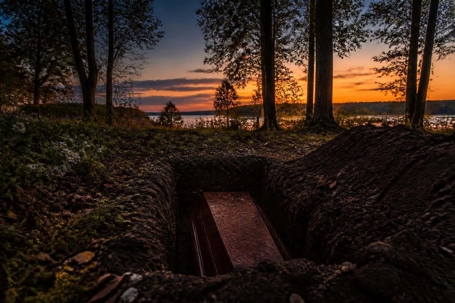 Uma cova aberta em um cenário natural ao entardecer, cercada por árvores e folhas caídas, transmitindo uma atmosfera de reflexão e mistério.